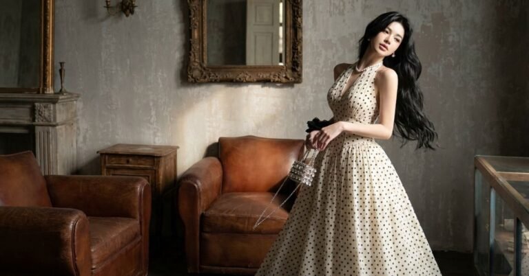 Ultra-realistic DSLR-style Google Discover feature image featuring SAME female model from reference image with EXACT SAME face, SAME long black silky hair, SAME slim feminine body proportions, SAME elegant luxury aura. She stands gracefully inside a luxurious vintage-inspired mansion room with textured grey walls, warm cinematic lighting, and elegant old-money interior décor. Model positioned on RIGHT side of frame wearing a flowing ivory-white polka dot maxi dress with soft black polka patterns and elegant movement in fabric. She holds a luxury crystal evening handbag while softly turning her body naturally. Soft glamorous makeup, delicate pearl jewelry, and elegant feminine heels complete the look. LEFT side contains large clean negative space for bold Discover-style headline text overlay. Old money feminine luxury aesthetic, French-inspired fashion vibe, cinematic warm shadows, soft romantic atmosphere, ultra realistic skin texture, shallow depth of field, 85mm DSLR lens effect, ultra HD quality, no blur, no watermark.